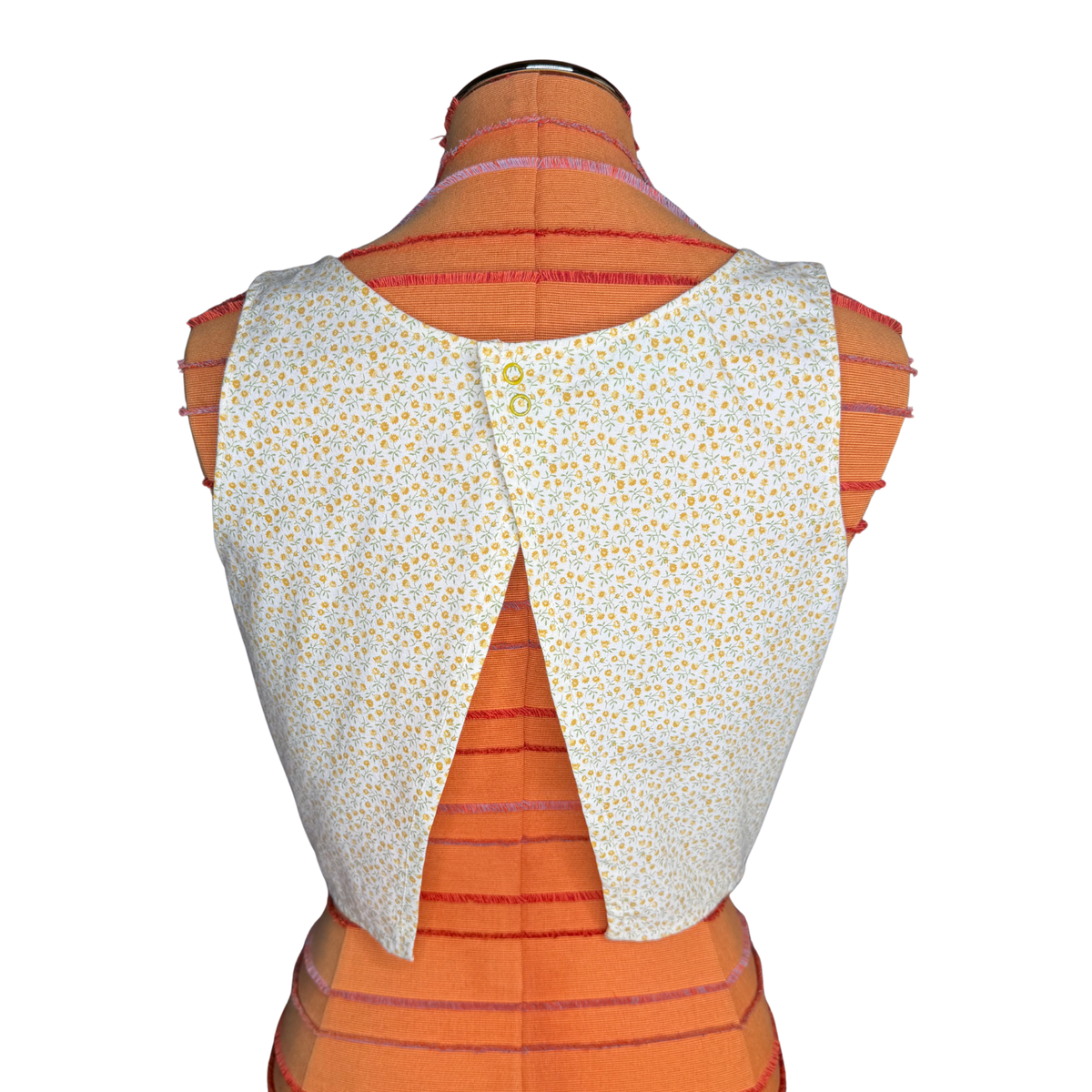 Back view of a sleeveless vest or cape-style top with a white base and small yellow floral print, displayed on an orange dress form against a white background.