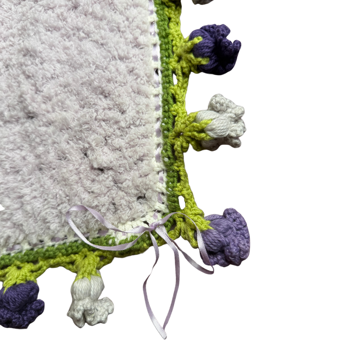 Close-up of a handmade crocheted textile corner with a light gray fuzzy interior, lime green crochet edging, lavender and white pom-pom details, and a small lavender ribbon bow on a white background.
