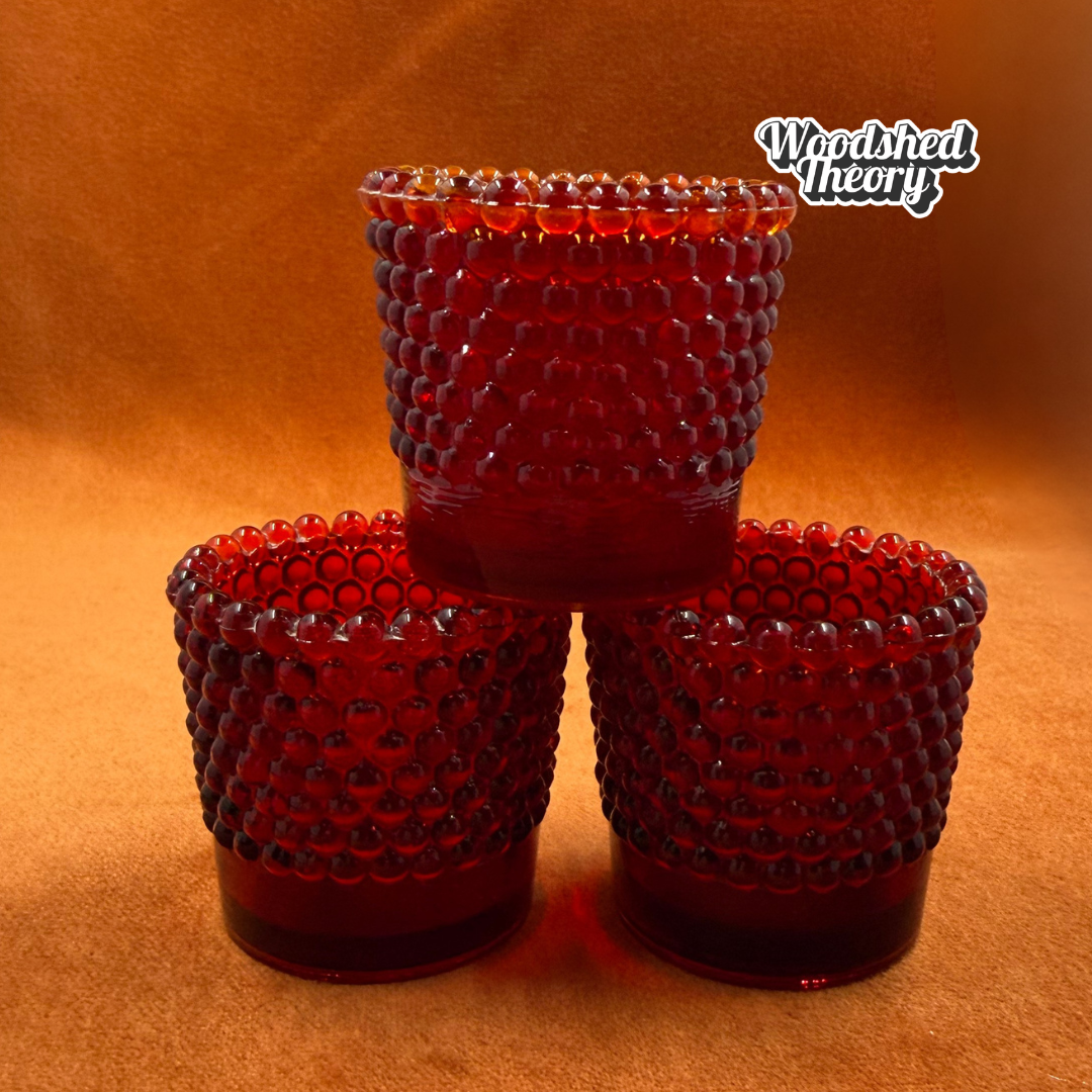 Three vintage-style red glass candleholders with a textured, beaded bubble pattern, arranged in a pyramid stack on a warm brown background. A 'Woodshed Theory' brand logo is visible in the top right corner.