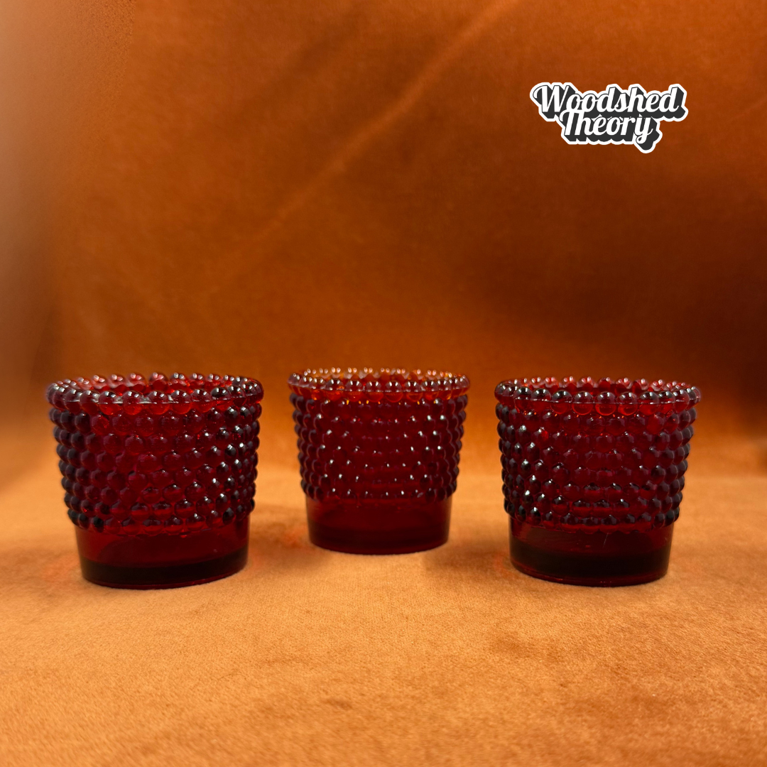 Three small dark red beaded glass shot glasses arranged in a row against a warm brown textured background, with the 'Woodshed Theory' logo in the top right corner.