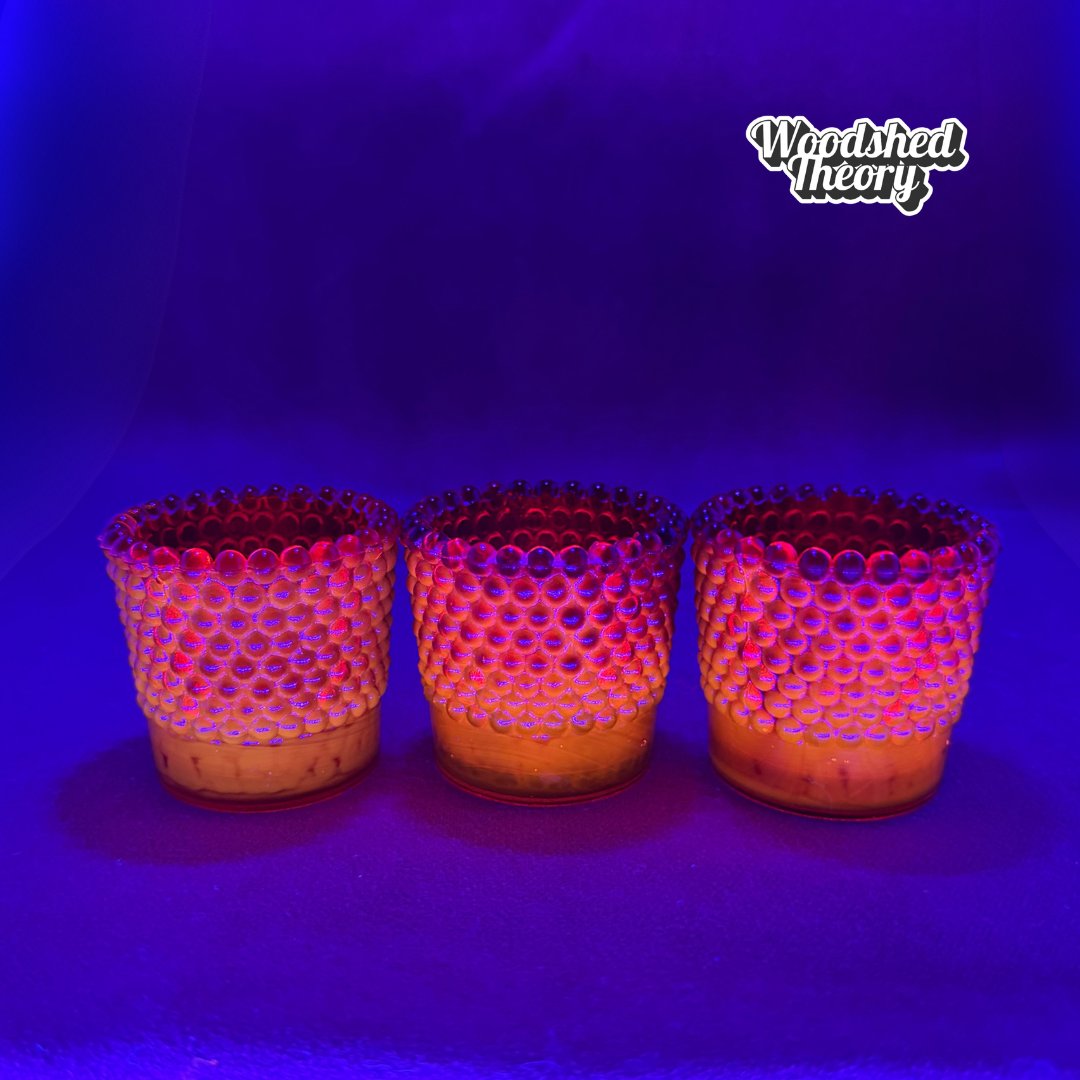 Three small textured glass candle holders with a honeycomb pattern, glowing with pink and orange light under a purple/blue UV lamp. The 'Woodshed Theory' logo is visible in the top right corner.