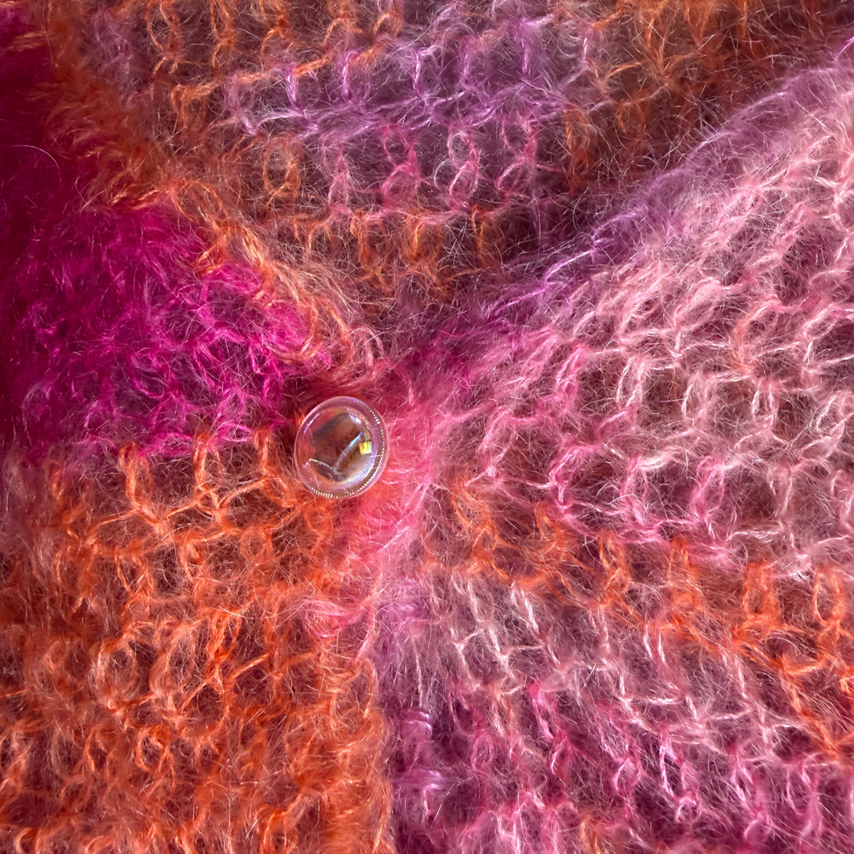 A close-up of a fuzzy, textured knit fabric in a variegated blend of bright pink, magenta, orange, and mauve, with a small clear iridescent button resting near the center.