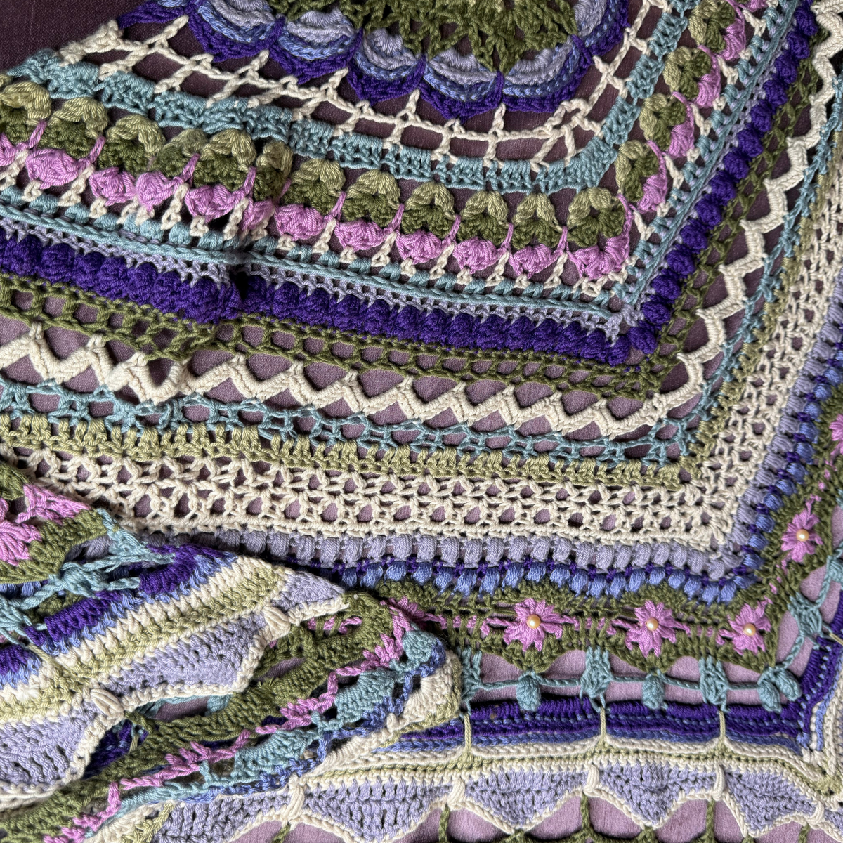 A close-up of a handmade crocheted granny square blanket featuring intricate patterns in purple, olive green, light blue, and cream, with small pink floral accents.