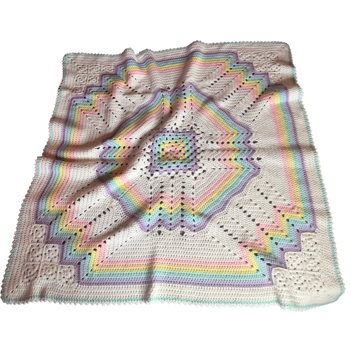 A handmade crocheted blanket with a geometric starburst pattern in pastel rainbow colors on a cream base, laid flat against a white background.