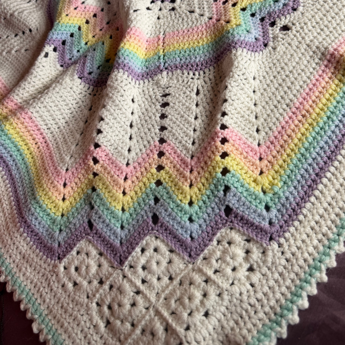 A close-up of a handmade crocheted blanket featuring a cream base with a pastel rainbow pattern and chevron-like zigzag stripes.