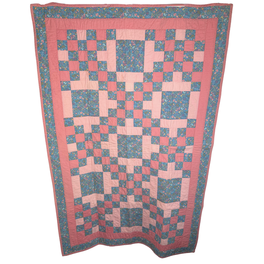 A vintage-style patchwork quilt hanging flat against a white background. The quilt features a geometric block pattern in shades of pink and blue floral print, framed by a solid pink border.