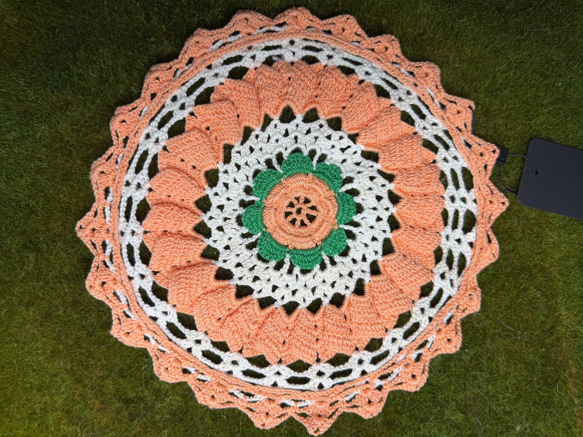A crocheted round doily with a scalloped edge, featuring concentric rings of peach, white, and green crochet patterns, laid flat on green grass next to a black tag. back