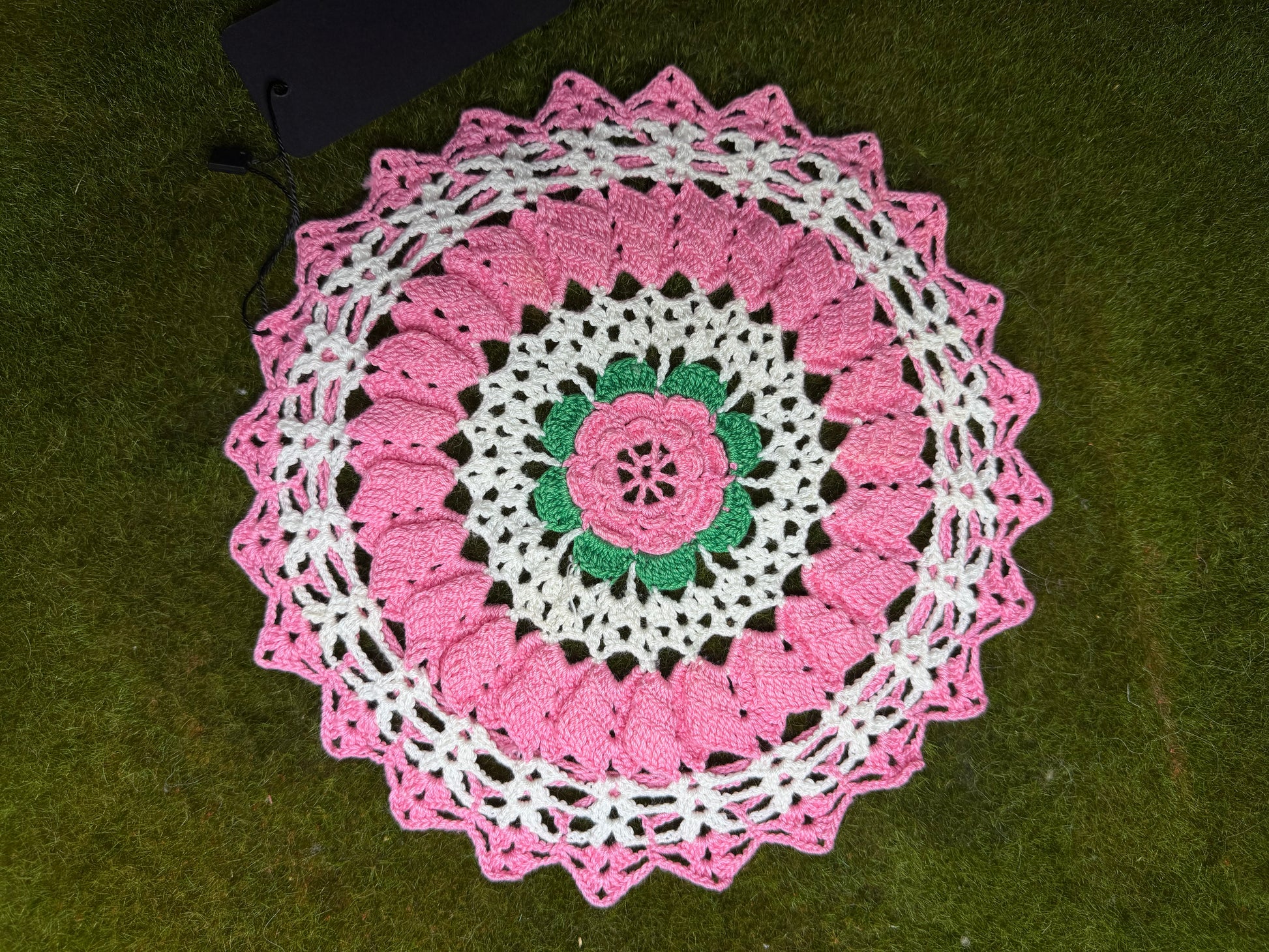 A handmade crocheted doily with a pink and white floral mandala design and a green center, laid flat on green grass.