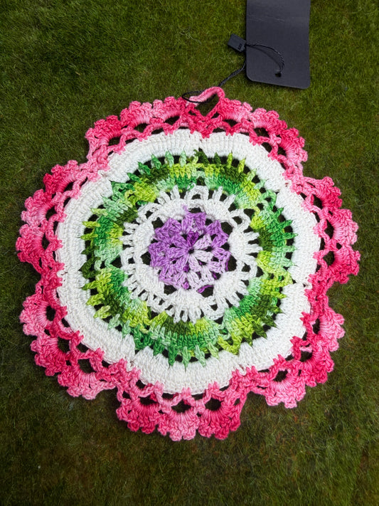 A circular crocheted doily with concentric rings of purple, green, white, and pink, laid flat on green grass next to a black tag.