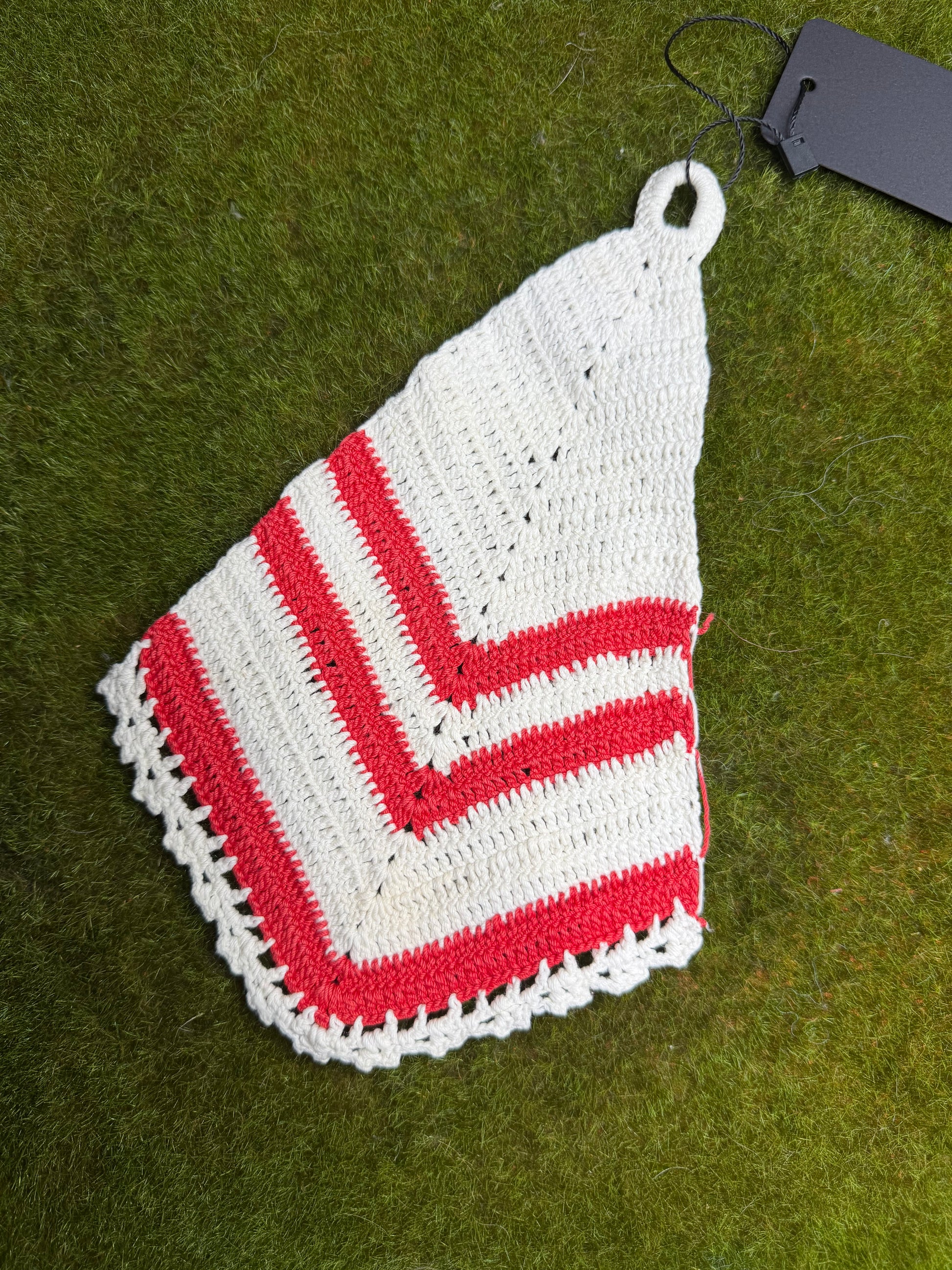 A handmade crocheted potholder in white and red, laid flat on green grass with a black hangtag attached.