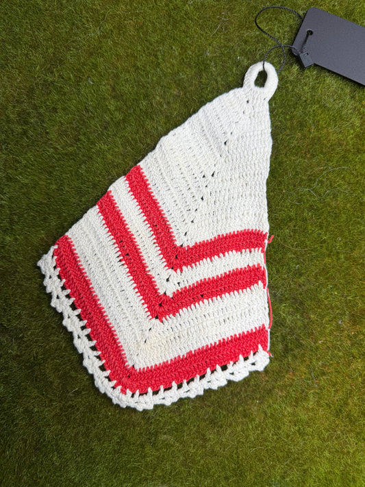 A handmade crocheted potholder in white and red, laid flat on green grass with a black hangtag attached.