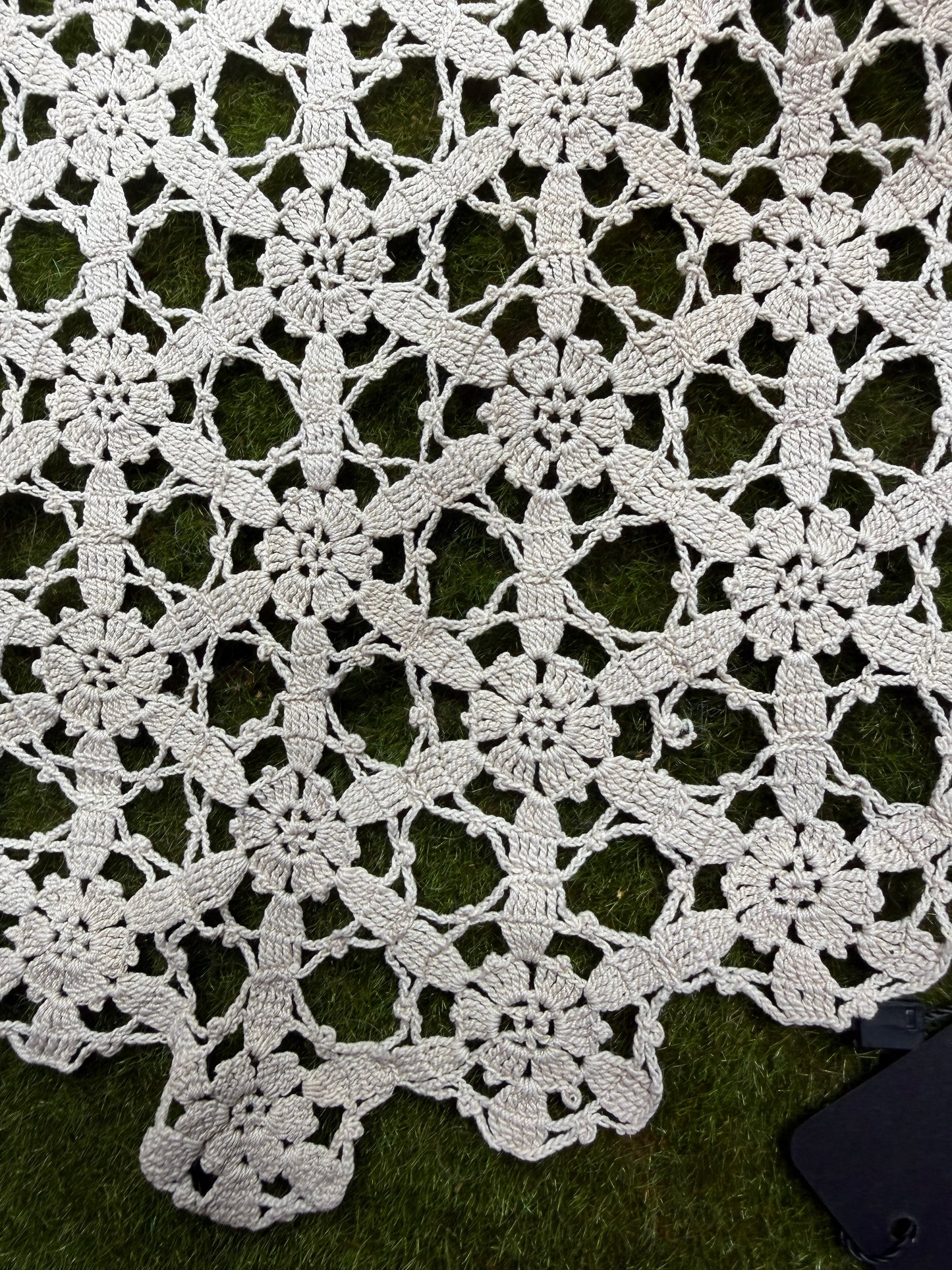 A close-up of a white crocheted lace textile with an intricate floral and geometric openwork pattern, laid flat on a green grass-like surface.
