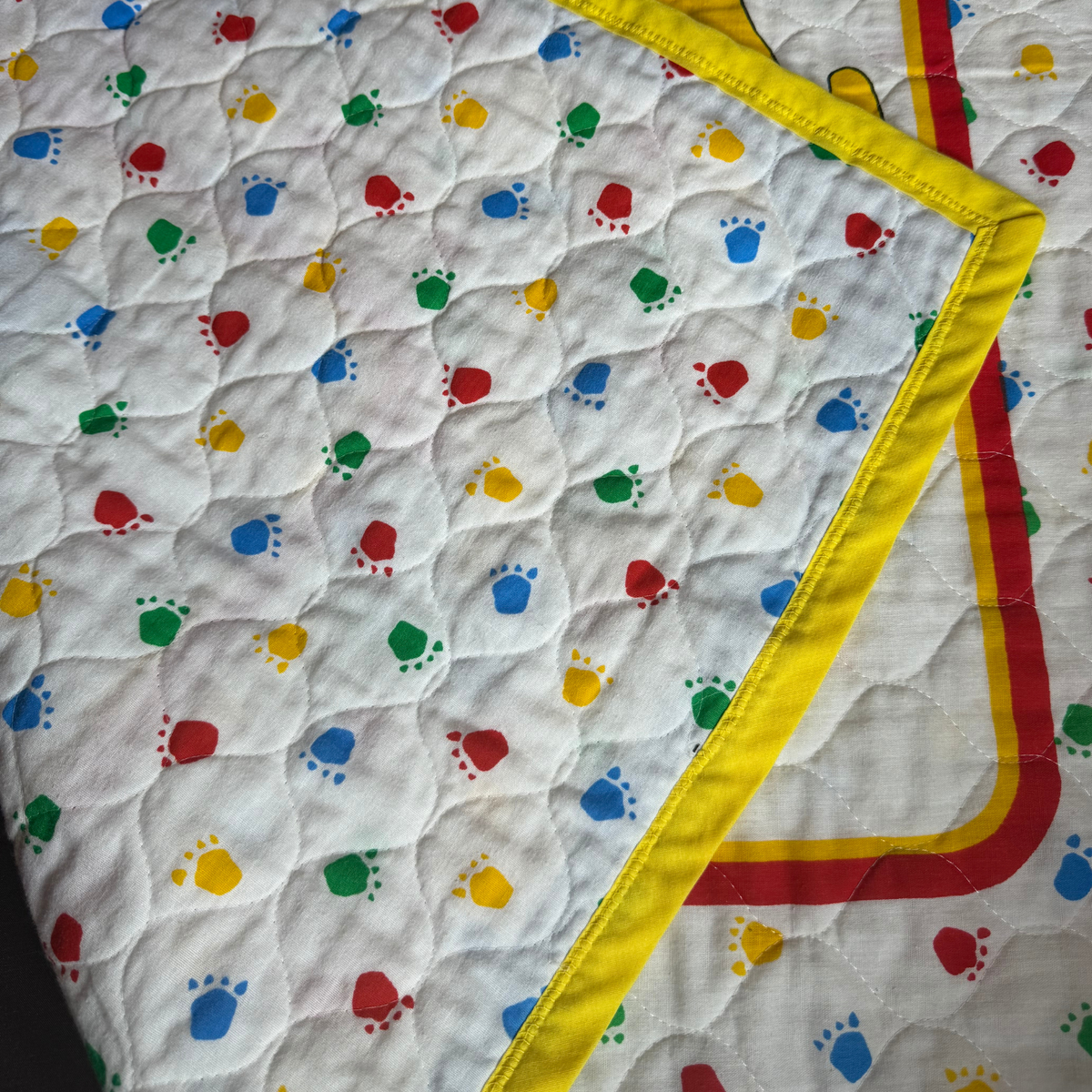 A close-up of a quilted baby blanket or quilt with a white background, colorful printed paw prints in red, blue, green, and yellow, and a yellow border with red trim.