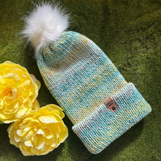 A beanie with a green, blue, and yellow color pattern, featuring a white faux fur pom-pom, a custom cork tag, and displayed next to two yellow flowers on a green surface. garden stroll front